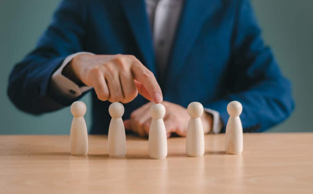 Businessperson selecting one of five wooden figures arranged in a row on a desk.