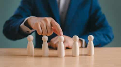 Businessperson selecting one of five wooden figures arranged in a row on a desk.