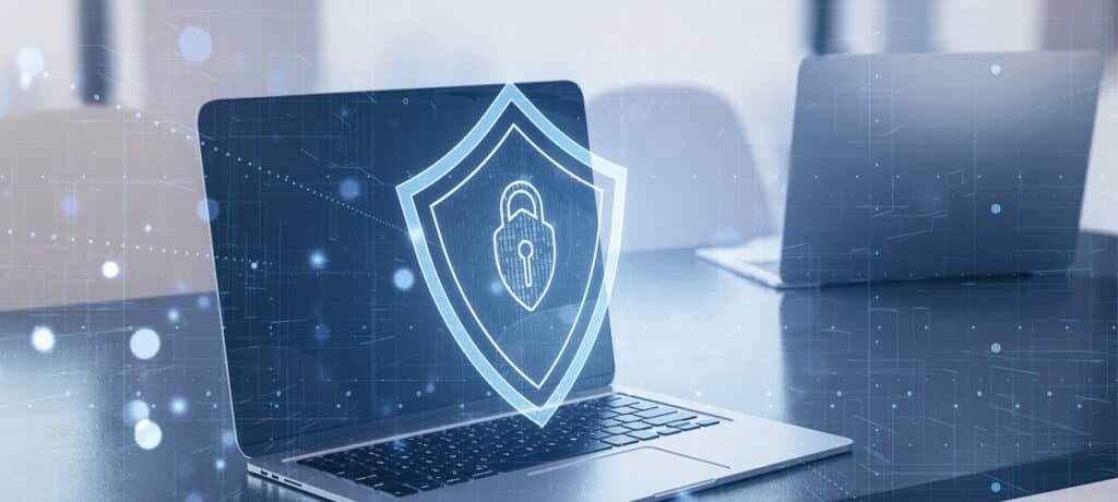 Laptop with a digital shield symbol onscreen signifies cybersecurity concept, blended with an office interior background. Explore what NIST Cybersecurity Framework is and what it means